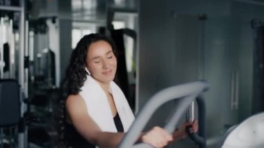 Neşeli spor antrenmanı. Genç, sağlıklı bir kadın spor salonunda eliptik eğitim aparatları üzerinde egzersiz yapıyor ve kablosuz kulaklıkla müzik dinliyor, antrenmanların keyfini çıkarıyor.