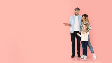 Aile teklifi. Neşeli Orta Çağ Ebeveynleri ve Küçük Kız, Pembe Stüdyo 'nun arka planında parmak gösterip, boşluğa sarılırken poz veriyor. Şuraya bak konsept. Panorama