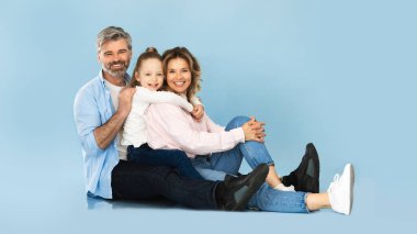 Üç kişilik mutlu bir aile. Orta yaşlı ebeveynler ve küçük kızı oturmuş mavi arka planda kameraya gülümsüyor. Anne, baba ve çocuğun birlikte poz verirken çekilmiş bir stüdyo fotoğrafı. Panorama