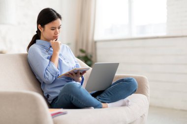 Konsantre olmuş genç Çinli bayan online eğitim dersine katılıyor, kucağında bilgisayarla koltukta oturuyor, dizüstü bilgisayarı tutuyor, dizüstü bilgisayara bakıyor, evden ders çalışıyor, boşluğu kopyalıyor.