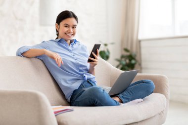 İnternette iş arayan Asyalı genç bir kadın, rahat oturma odasında koltukta oturuyor, yepyeni bir akıllı telefon ve dizüstü bilgisayar kullanıyor, internetteki kariyer fırsatlarını kontrol ediyor, kameraya gülümsüyor, uzay boşluğunu kopyalıyor.