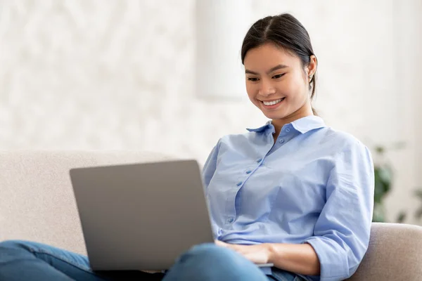 穿着休闲装的年轻漂亮的亚洲女人坐在家里的沙发上 使用现代笔记本电脑 在电脑键盘上打字 在约会网站上与男人聊天 复制空间 — 图库照片
