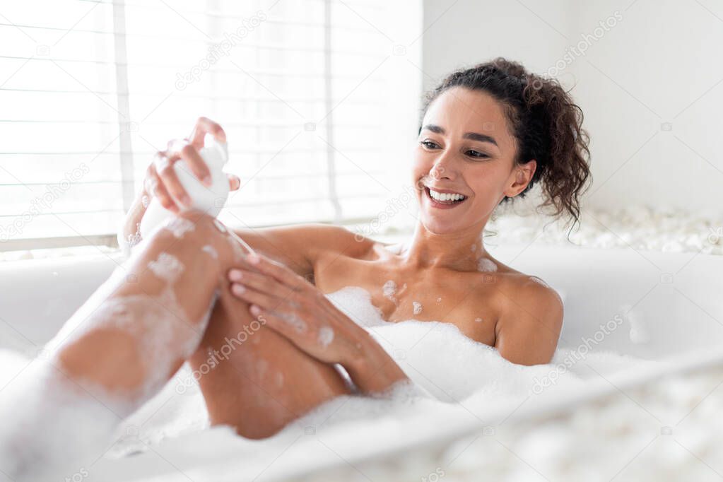 Pretty young woman applying soap on her body while lying in hot bubbly bath at home. Cheerful millennial lady enjoying relaxing spa day in bathtub near window at luxury hotel