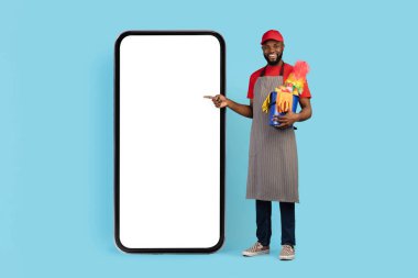 Ev Temizliği Hizmetleri Uygulaması. Siyah, yakışıklı, önlük takan, beyaz ekranlı, büyük, boş, akıllı telefonu işaret eden, elinde sepet dolusu deterjan tutan, gülümseyen, mavi arka plan,