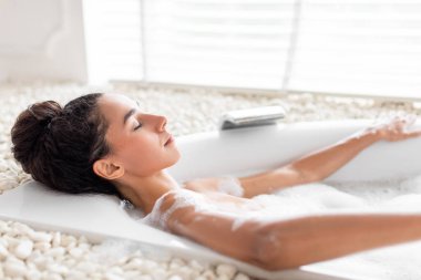 Sexy young woman lying in bathtub with closed eyes, enjoying beauty ritual at domestic spa, free space. Lovely millennial lady resting in hot relaxing bath at home. Wellness concept clipart