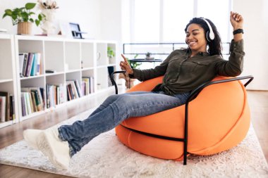 Aşırı mutlu siyah kadın, kablosuz kulaklık takıyor oturma odasında puf koltukta dans ediyor, popüler müzik dinleyen, en sevdiği şarkıya yönelen, tüm vücut uzunluğuyla mutlu, genç bayan.