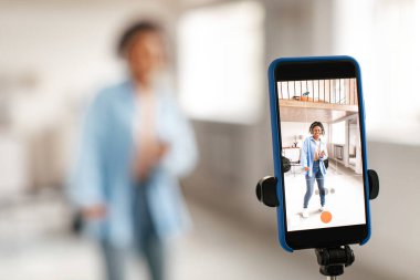 Sosyal Medya Yarışması. Kulaklıklı neşeli siyahi kadın kamerada dans ediyor evde cep telefonunu tripodla kullanıyor, cep telefonuyla internete yüklemek için son moda içerik yaratıyor, ekrana odaklanıyor.