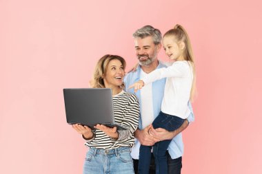 Neşeli Aile Laptop Bilgisayar Reklam Sitesi ile Pembe Arkaplanda Duruyor. Ebeveynlerin Stüdyo Çekimi ve Küçük Kızın İnternete Birlikte Bakması. Teknoloji Konsepti