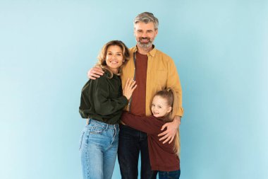 Orta yaşlı ebeveynler ve küçük kız mavi arka planda birbirlerine poz veriyorlar, kameraya gülümsüyorlar. Stüdyoda ayakta duran üç kişilik mutlu bir aile fotoğrafı.