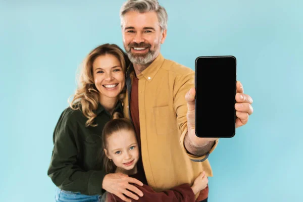 Mobile Offer. Family Holding Cellphone Showing Empty Screen To Camera Posing Over Blue Background In Studio. Cheerful Parents And Little Daughter Advertising Application. Selective Focus On Phone