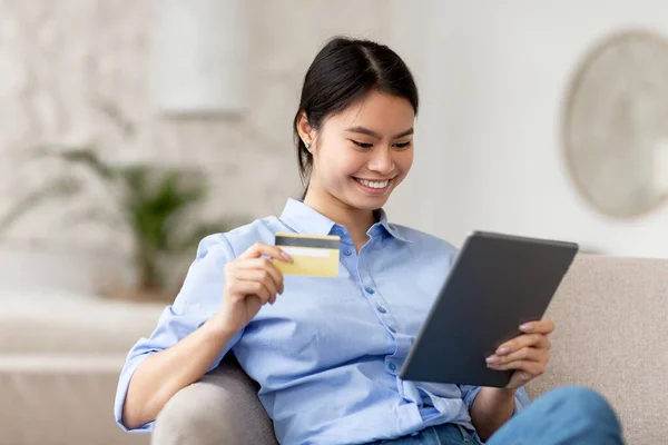 在家里的沙发上 年轻快乐的日本女人穿着休闲装 拿着现代数码平板电脑 上网购物 用黄色信用卡 复制空间 网上银行 金融移动应用 — 图库照片