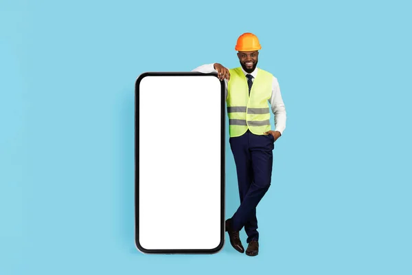 Mobile Ad. Handsome Black Civil Engineer Standing Near Big Blank Smartphone With White Screen, Smiling African American Builder Wearing Safety Vest And Hard Hat Recommending New Application, Mockup