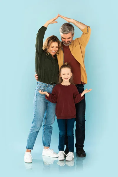 Family Housing. Vertical Shot Of Parents Making Home Roof Above Their Daughters Head Joining Arms Posing Standing Over Blue Studio Background. Real Estate Offer Concept