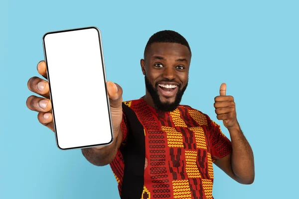 伝統的な衣装を着た陽気なアフリカ人男性は 空白のスマートフォンを保持し 親指を表示します ホワイトスクリーンで携帯電話を実証興奮黒人男性 新しいアプリをお勧めします コラージュ モックアップ — ストック写真