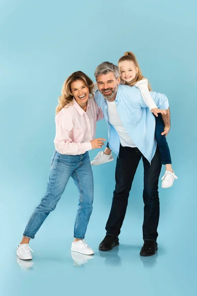 Vertical Shot Of Happy Family Posing Together, Daddy Carrying Daughter Piggyback Smiling To Camera On Blue Studio Background. Joy Of Parenthood Concept. Full Length