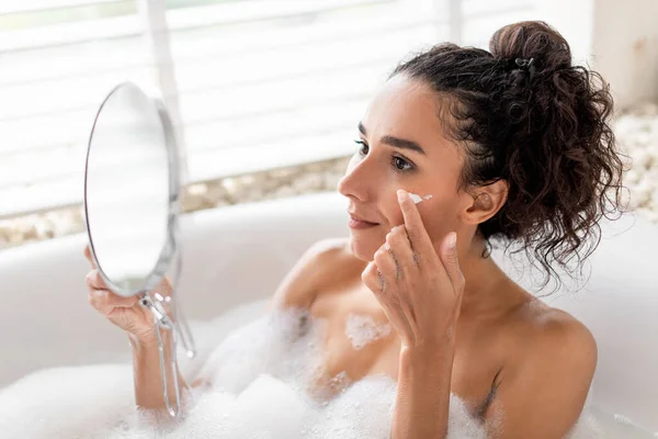 快乐的千禧年女性在浴缸里使用面部保湿霜 在家里照镜子 快乐的年轻女子一边在室内泡泡浴一边在脸上涂营养霜 — 图库照片
