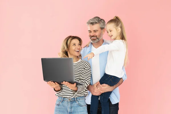 Joyful Family Using Laptop Computer Advertising Website Standing Over Pink Background. Studio Shot Of Parents And Little Daughter Browsing Internet Together. Technology Concept