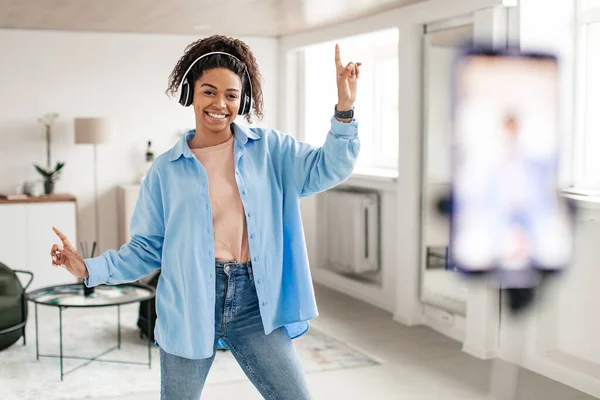 Joyful black woman in wireless headseat dancing at camera filming video using phone on tripod at home creating trendy content on mobile app to share on social media, selective focus on millennial lady