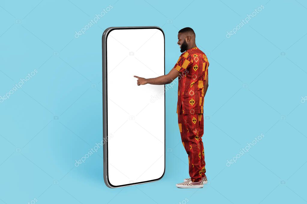 Great App. Happy African Man In Traditional Costume Touching White Screen Of Big Smartphone While Standing Isolated On Blue Background, Black Guy Showing Copy Space For Mobile Ad, Collage, Mockup
