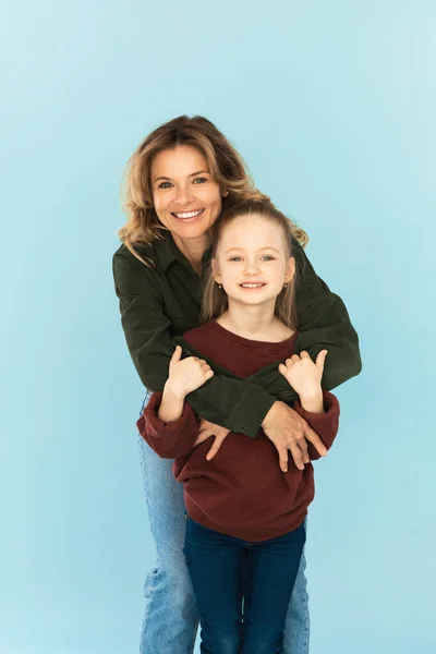 Cheerful Middle Aged Mother Embracing Her Little Daughter Standing Posing In Studio Over Blue Background, Smiling To Camera. Family, Joy Of Motherhood Concept. Vertical Shot