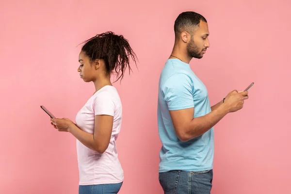 Social network, gadget addiction. Millennial black couple using mobile phones, standing back to back, ignoring each other, playing video games on pink studio background