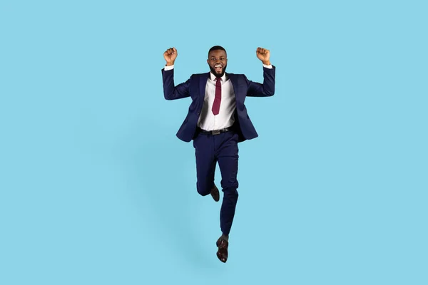 Business Win. Joyful Black Businessman In Suit Jumping With Clenched Fists Over Blue Studio Background, Excited African American Male Entrepreneur Emotionally Celebrating Success, Copy Space