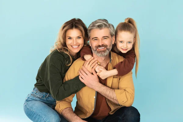 Happy Family Hugging Smiling To Camera Posing Over Blue Background, Studio Shot. Middle Aged Parents And Little Daughter Expressing Positive Emotions. Shot Of Mom And Kid Girl Embracing Dad