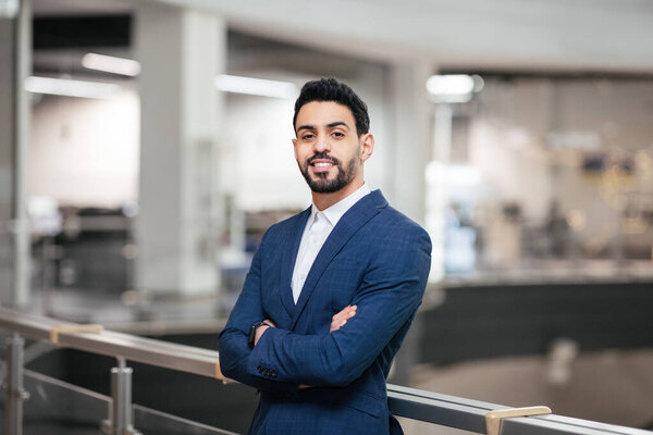 Smiling confident calm attractive young middle eastern businessman with beard in suit with crossed arms looks at camera in office interior. Successful business, ceo manager and leadership at factory