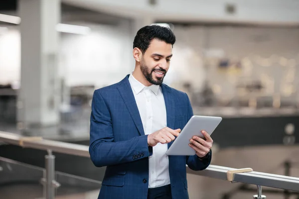 Cheerful confident young arab general manager with beard in suit works on tablet in office interior, control plant work. Modern online technology for successful business, ceo career and leadership