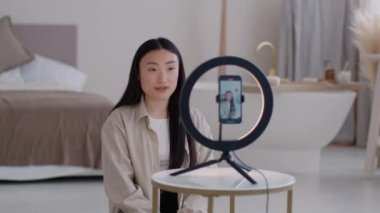 Yaşam tarzı blogu. Genç pozitif, güzel Asyalı blog yazarı sosyal medya blogu için konuşma videosu kaydediyor, akıllı telefon ve dairesel lambanın önünde oturuyor, seçici odaklanma, yavaş çekim