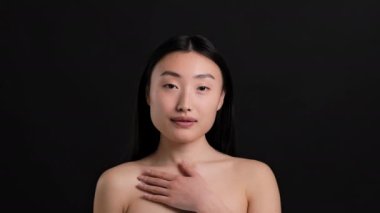 Vücut nemlendirici konsept. Genç, güzel Koreli bir kadın, göğsünde krem kalple poz veriyor, kameraya krem sürüyor ve gülümsüyor, siyah stüdyo arka planı, yavaş çekim.