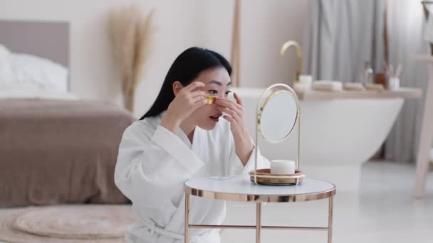 乌鸦脚的预防 年轻美丽的亚裔女士在眼皮底下涂上金色胶原蛋白片 早上坐在浴室里 看着镜子 慢动作 空旷的空间 — 图库视频影像