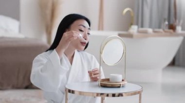 Cilt bakımı ve vitamizasyon. Genç, güzel Asyalı bir bayan aynaya bakarak besleyici serum uyguluyor, evde yüz şımartma ayinleri yapıyor, yavaş çekim, serbest alan.