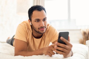 Orta Doğulu adam kulaklık takıyor, cep telefonu kullanıyor interneti tarıyor ve evdeki modern yatak odasında yalan söylüyor. Gadgets Yaşam Tarzı Konsepti. Seçici Odaklanma