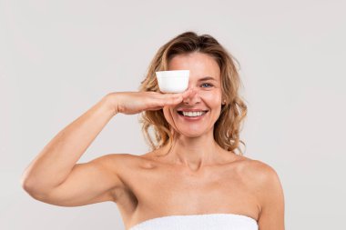 Anti-Age Kozmetik. Havluya sarılmış, avucunda nemlendirici kremle gülümseyen orta yaşlı bir kadının portresi bir gözünü yeni cilt ürünüyle kaplıyor ve gri arka planda poz veriyor.