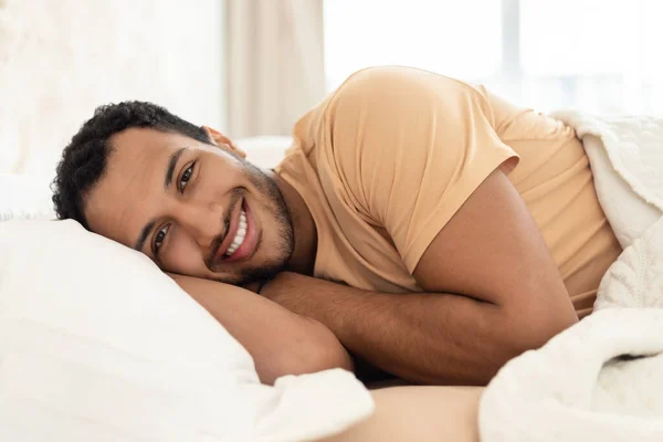 カメラに向かって微笑むベッドで寝そべっている陽気なアラビア人の男が 自宅のモダンなベッドルームで休息とリラクゼーションのために休息の頭を置いています レクリエーションと国内快適コンセプト — ストック写真