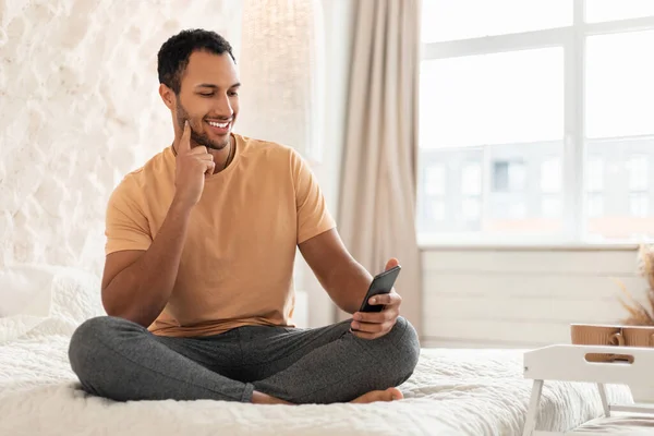 Happy Arabic Male Texting Using Phone Sitting On Bed, Reading News Online And Using Mobile Application In Modern Bedroom At Home. New App Advertisement, Technology And Gadgets