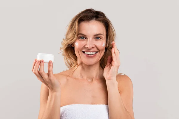 迷人的中年妇女使用保湿面霜 漂亮的成熟女性用抗老化化妆品包裹在毛巾罐中 在相机前微笑 复制空间 — 图库照片