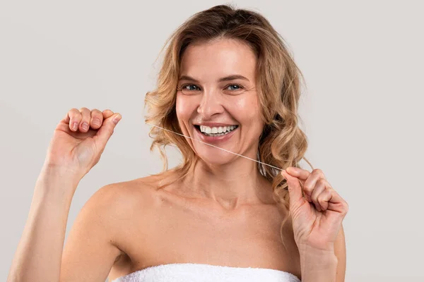 口腔衛生 歯科用フロスを使用した中年女性の笑顔とカメラを見て 魅力的な熟女は毎日の衛生を作ります ライトスタジオの背景にタオルで包まれて立ちます スペースをコピー — ストック写真
