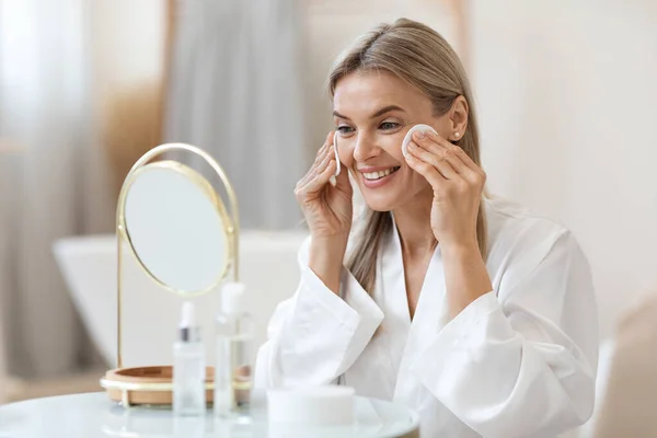 穿着白色浴衣的漂亮女士正坐在浴室的镜子前 擦去脸上的化妆品 用棉垫和清洁剂 侧视图 特写照片 — 图库照片