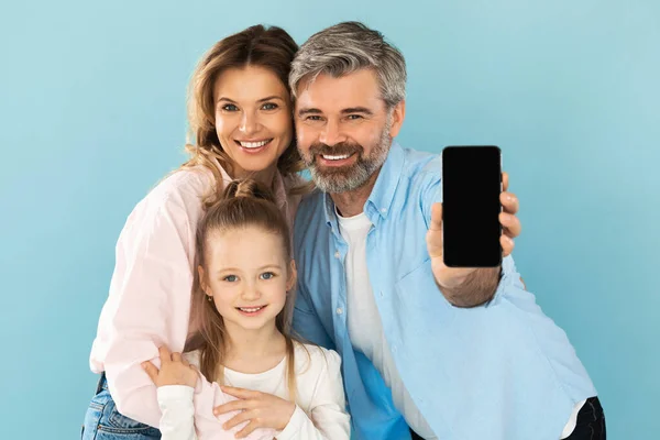 Mobile Offer. Family Showing Smartphone With Blank Screen Posing Embracing Smiling To Camera Standing On Blue Background In Studio. Great Application Advertisement. Mockup