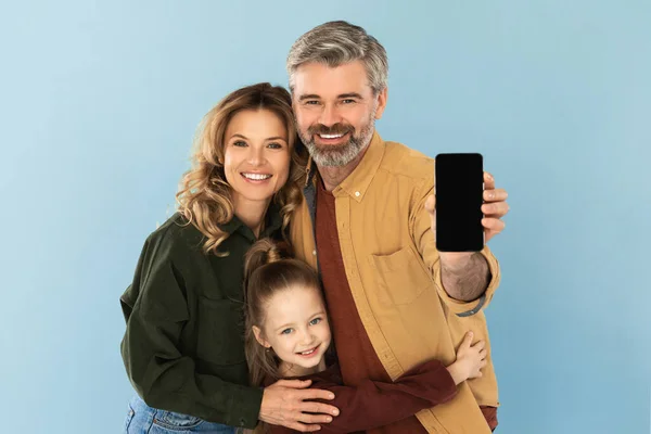 Great Application. Family Holding Mobile Phone Showing Blank Screen To Camera Recommending App Posing In Studio On Blue Background. Parents And Daughter Advertising Offer Via Smartphone. Mockup