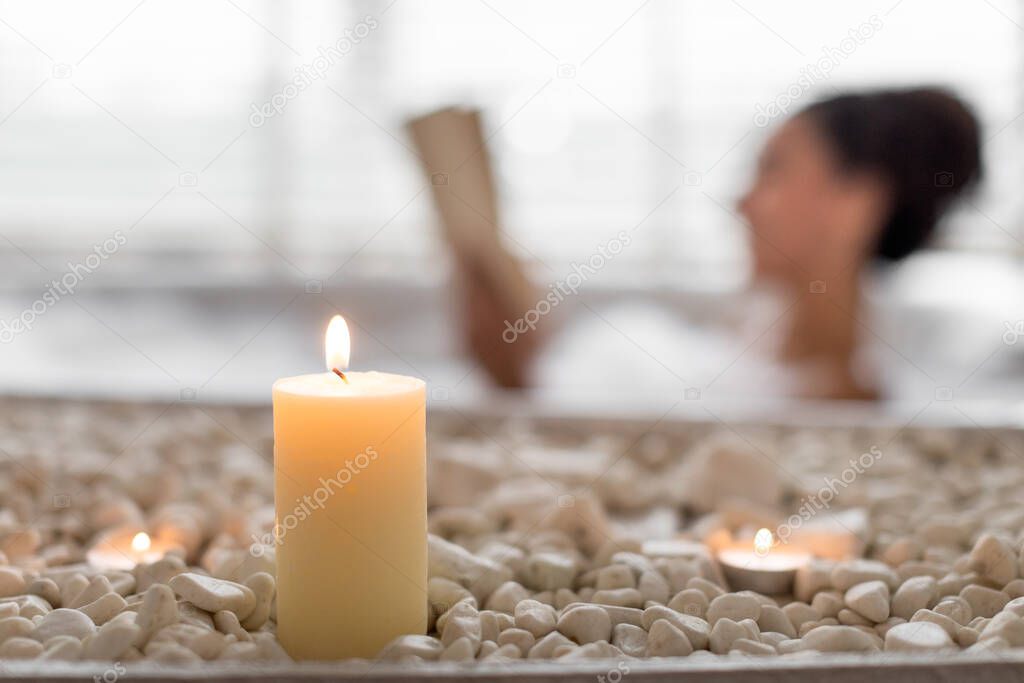 Young woman lying in foamy bath, reading book in relaxing atmosphere, selective focus on candles and stones. Millennial female enjoying interesting story in bathtub at home, free space