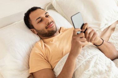 Orta Doğulu Adam, yatak odasında yatakta uzanan sosyal medyada Smartphone Networking Sting kullanıyor. Adamın evdeki mobil uygulamayı kullanırken yüksek açılı görüntüsü. Teknoloji ve Malzemeler