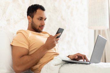 Diz üstü bilgisayar kullanan, cep telefonu kullanan, internette mesajlaşan ve evde yatağında oturan Arap serbest çalışan adam. Ciddi Adam Akıllı Telefon 'da İnternet Taraması Uygulaması Kullanıyor. Uzaktan kumanda. Yan Görünüm
