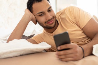 Orta Doğulu adam cep telefonu kullanıyor. İnternetten mesajlaşıyor. Evde yatakta yatıyor. Erkek, yeni uygulamayı cep telefonuyla kullanıyor. İletişim, Teknoloji ve Gadget Kavramı. Seçici Odaklanma