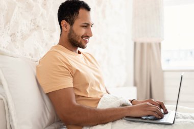 Çevrimiçi çalışan dizüstü bilgisayar kullanan ve evdeki yatak odasında oturan mutlu Arapça erkek serbest görüşü. Serbest ve Uzak Görev