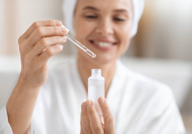 İpek bornozlu mutlu, sarışın kadın nemlendirici yüz serumu kullanıyor, güzellik ürünü taşıyor ve gülümsüyor, yakın çekim, kopyalama alanı. Yaşlanma karşıtı yüz bakımı konsepti