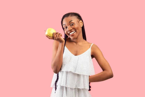 ピンク色のスタジオでリンゴを持っているかなり若いアフリカ系アメリカ人女性 カメラで笑顔でおいしいフルーツと陽気な千年黒の女性 健康な歯と美しい肌のためのビタミン — ストック写真