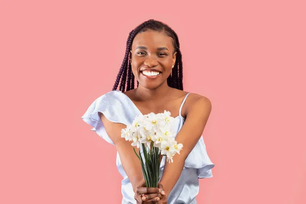 Happy millennial black lady holding bunch of daffodils over pink studio background. Positive millennial African American woman with bouquet of narcissus flowers celebrating birthday